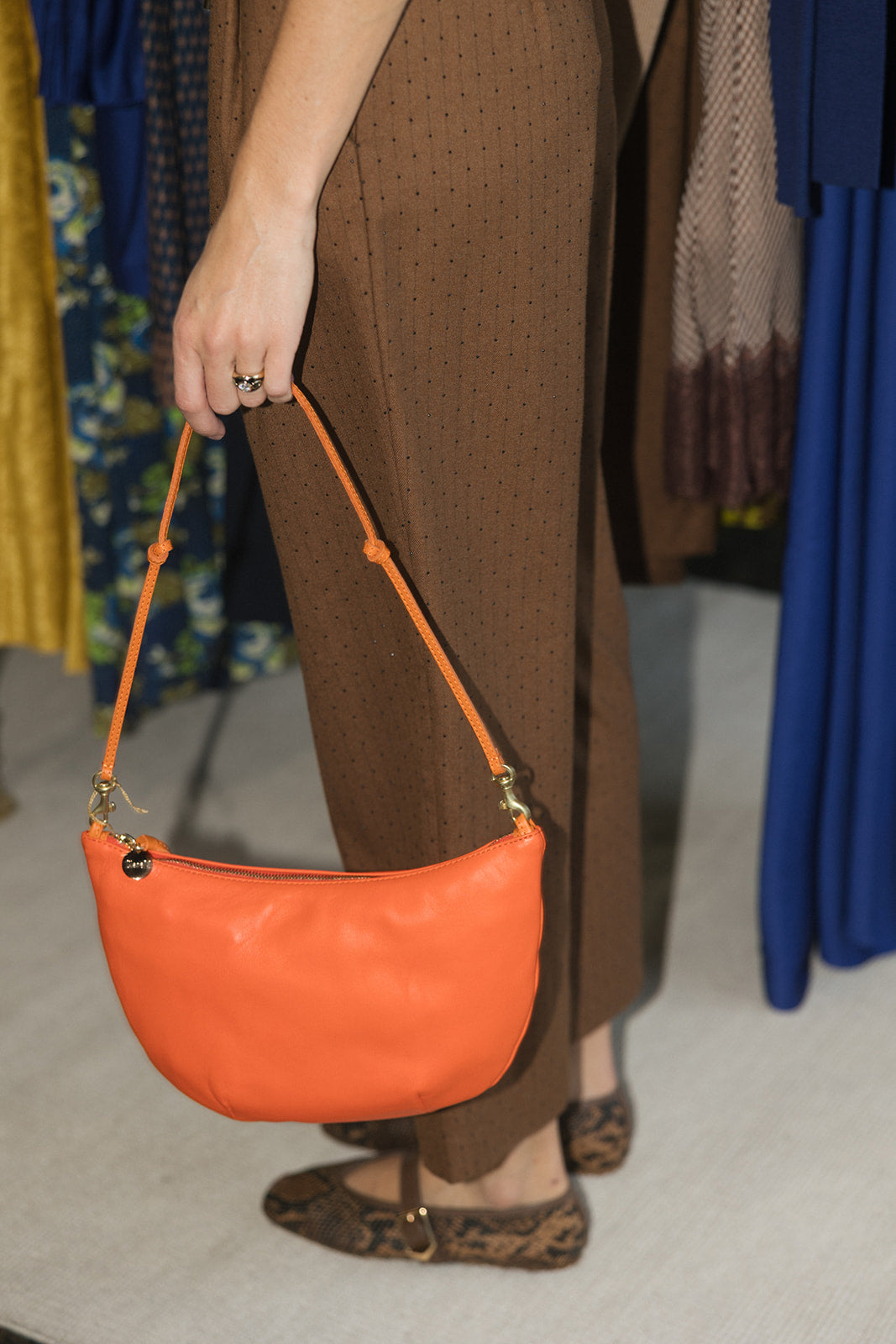 A person wearing brown patterned trousers and leopard-print flats holds a bright orange crescent-shaped leather shoulder bag with a matching strap in a clothing store.