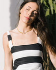 Woman wearing a black and white striped dress with a gold necklace, standing against a light background with plants.