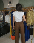 A woman viewed from behind wearing a white short-sleeve blouse and brown wide-leg trousers, standing in a boutique next to clothing racks.