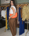 A woman posing in a clothing boutique, wearing a white sleeveless top, brown pleated trousers, and an orange shoulder bag, standing in front of a gold clothing rack.