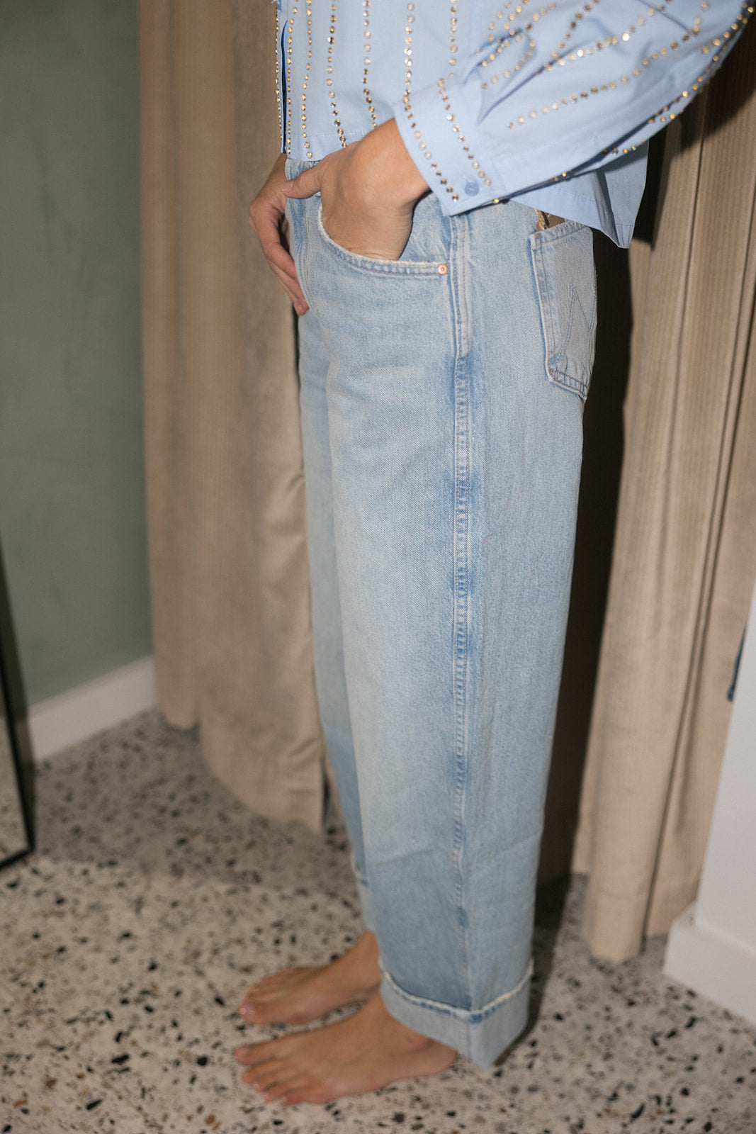 A person wearing light-wash blue denim jeans with a light blue embellished shirt, standing barefoot in an indoor setting with curtains and a speckled floor.