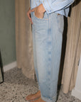 A person wearing light-wash blue denim jeans with a light blue embellished shirt, standing barefoot in an indoor setting with curtains and a speckled floor.