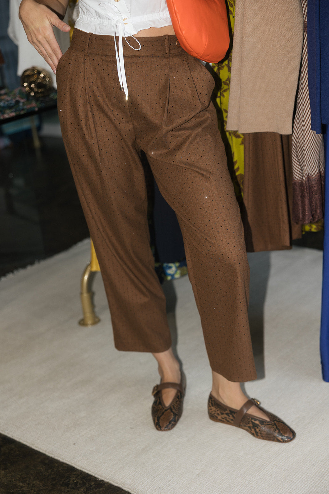 A person wearing brown cropped trousers with a white drawstring waist, styled with brown snakeskin Mary Jane flats and an orange shoulder bag in an indoor boutique setting.