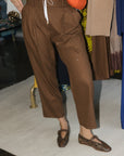 A person wearing brown cropped trousers with a white drawstring waist, styled with brown snakeskin Mary Jane flats and an orange shoulder bag in an indoor boutique setting.