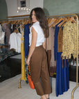 A woman stands in a boutique wearing a white sleeveless top and brown cropped trousers, holding an orange handbag. Clothing racks with blue, yellow, and brown garments are visible behind her.