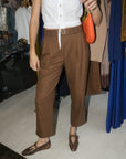 A person wearing brown pleated cropped trousers with a white sleeveless top and brown patterned flats, holding an orange bag in a clothing store.