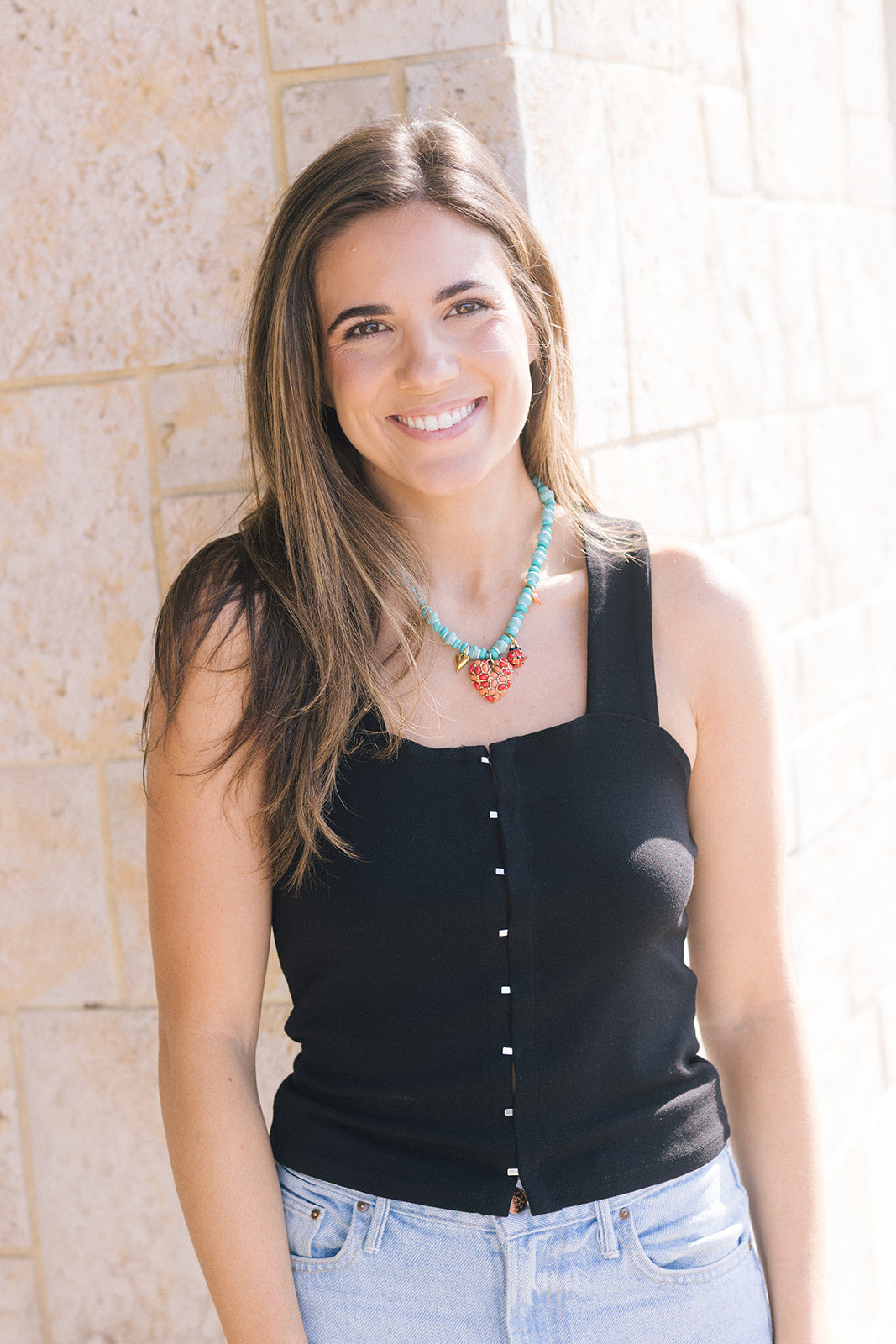 Woman wearing a black sleeveless top and light blue jeans with a necklace, standing against a stone wall.