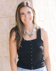 Woman wearing a black sleeveless top and light blue jeans with a necklace, standing against a stone wall.