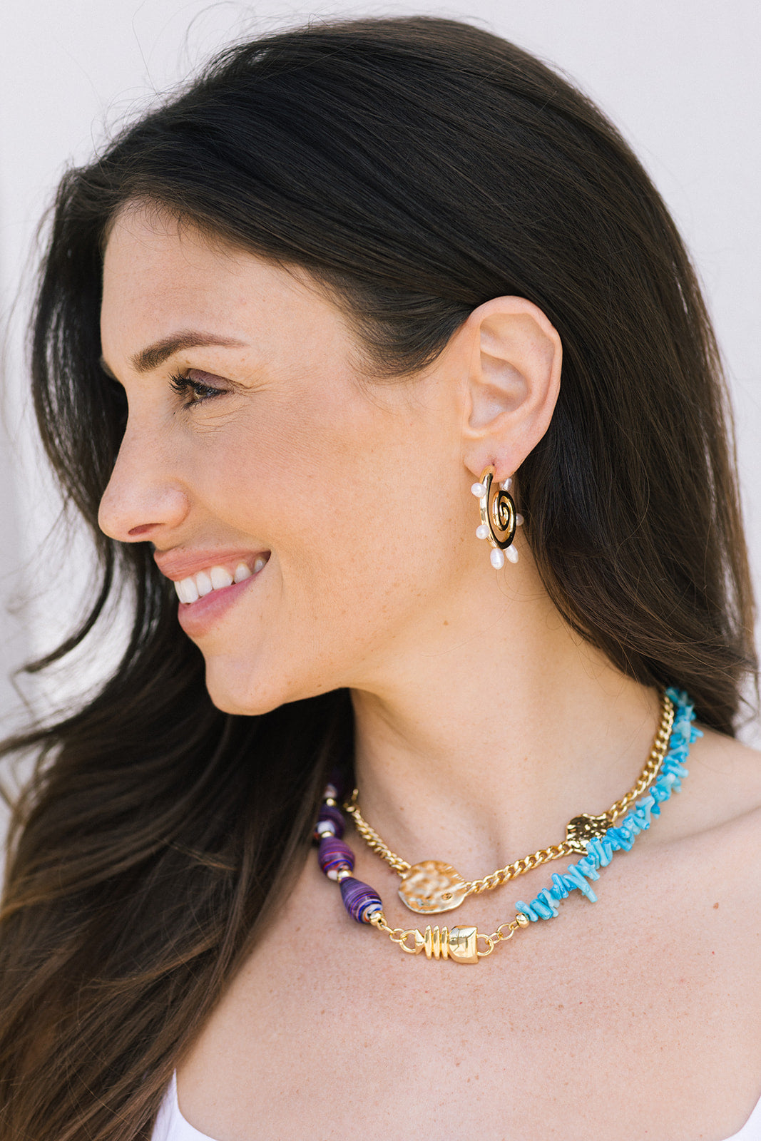 Woman wearing gold hoop earrings and colorful necklaces against a white background