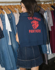 A woman wearing a navy blue cropped hoodie with a red 'THE GREAT PANTHERS' graphic on the back, standing in front of a clothing rack filled with jeans and other garments.