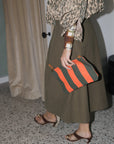 A person wearing a leopard-print top, an olive green skirt, and brown heeled sandals holds a striped orange and dark green clutch bag in an indoor setting with a speckled floor and beige curtains.