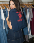 A woman wearing a navy blue cropped hoodie with a red 'THE ORLANDO PANTHERS' graphic on the back, paired with a denim mini skirt, standing in front of a clothing rack.
