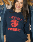 A woman smiling while wearing a navy blue crewneck sweatshirt with a red graphic that reads 'THE GREAT PANTHERS' and features a panther illustration. She is standing in front of a clothing rack filled with jeans.
