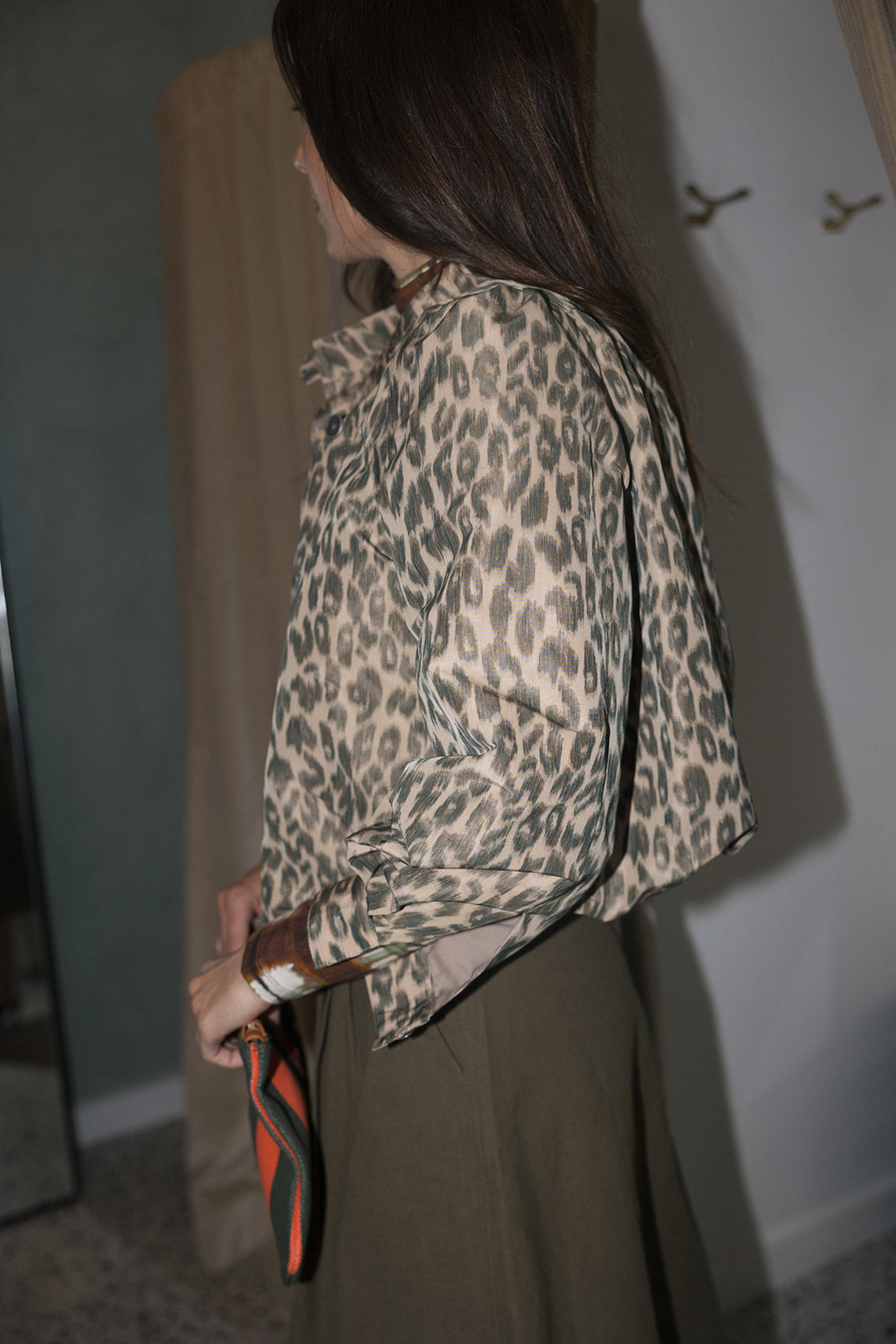 A woman wearing a beige and brown leopard-print long-sleeve blouse with rolled cuffs and an olive green skirt, standing in a fitting room with beige curtains., side