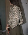 A woman wearing a beige and brown leopard-print long-sleeve blouse with rolled cuffs and an olive green skirt, standing in a fitting room with beige curtains., side