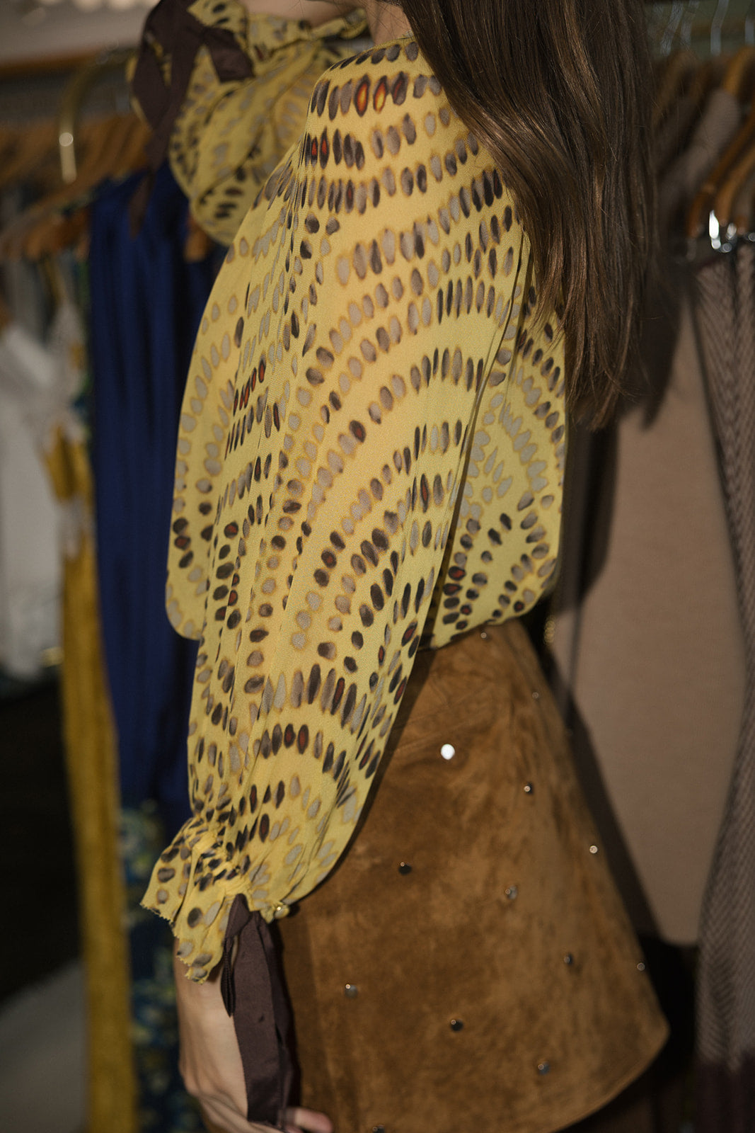 A close-up side view of a person wearing a yellow blouse with a brown abstract spotted pattern and a brown suede skirt with rhinestone embellishments, set against a background of clothing racks.