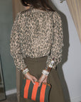 A woman wearing a beige and brown leopard-print long-sleeve blouse with rolled cuffs and an olive green skirt, standing in a fitting room with beige curtains, back