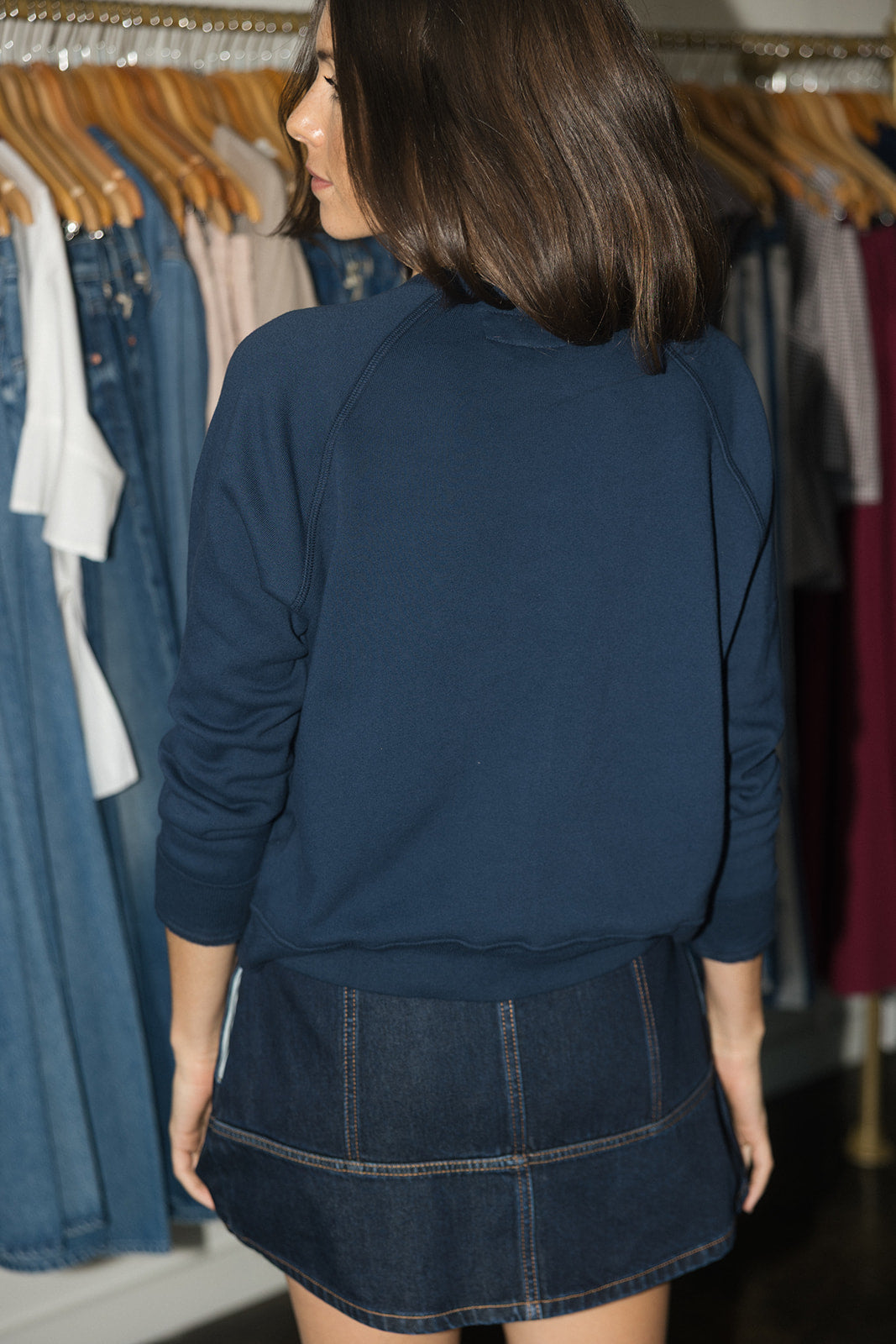 A woman shown from behind wearing a navy blue long-sleeve crewneck sweatshirt and a dark denim mini skirt, standing in front of a clothing rack.