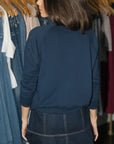 A woman shown from behind wearing a navy blue long-sleeve crewneck sweatshirt and a dark denim mini skirt, standing in front of a clothing rack.