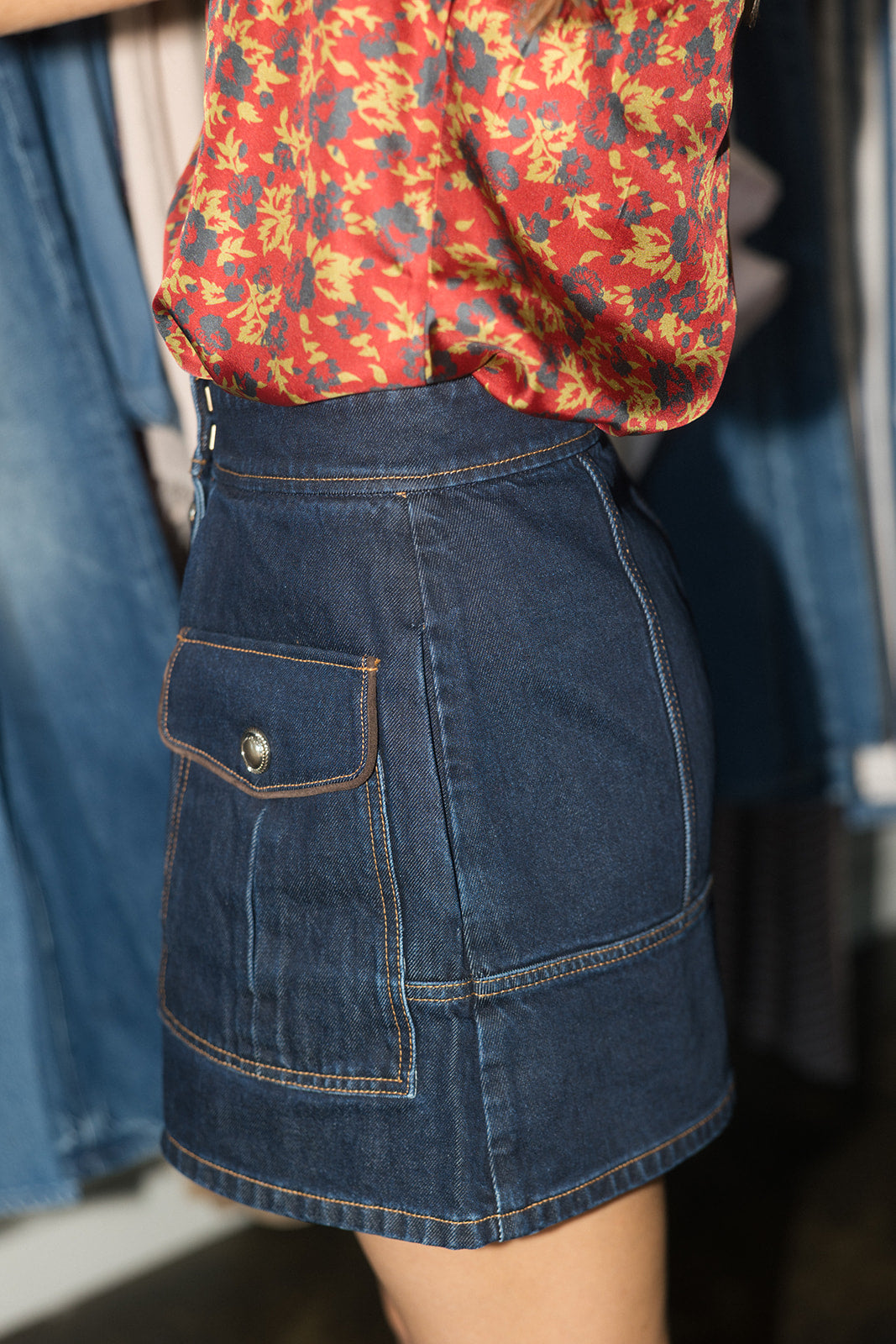 A close-up of a person wearing a dark blue denim mini skirt with a front flap pocket and contrast stitching, paired with a red floral blouse. The background shows a clothing rack with blue denim garments.