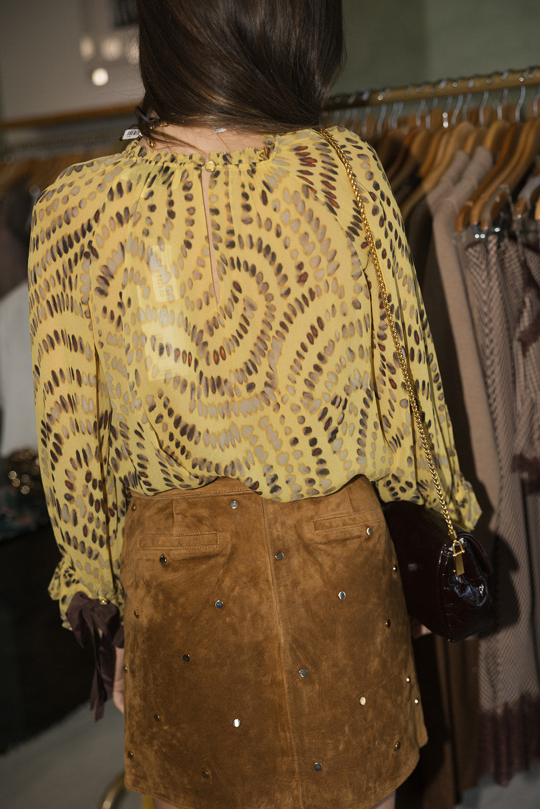 A person shown from behind wearing a sheer mustard yellow top with a brown abstract pattern and a brown suede skirt with rhinestone embellishments, standing in a clothing store.