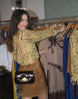 A woman with long brown hair stands in a clothing boutique, looking at garments on a rack. She is wearing a yellow open-knit long-sleeve top and a brown suede mini skirt, with a dark brown crossbody bag featuring a gold-tone clasp.
