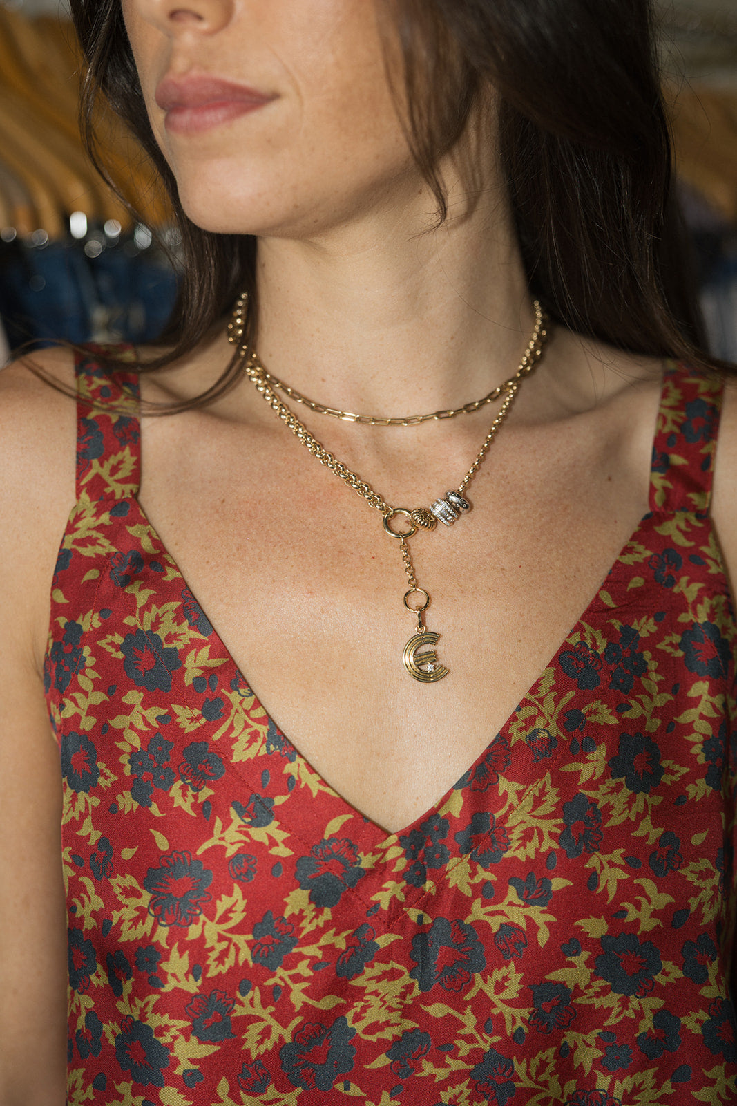 A close-up of a woman wearing a layered gold necklace with a small dangling charm, paired with a red and blue floral patterned top.