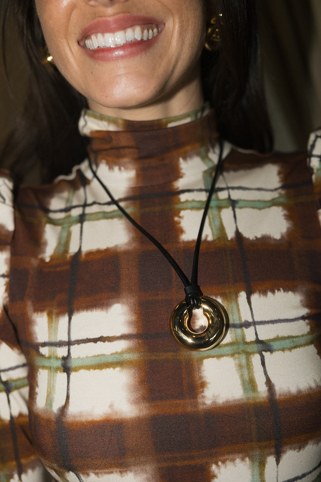 A close-up of a woman wearing a black cord necklace with a gold-toned circular pendant, paired with a brown and cream plaid turtleneck top.