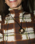 A close-up of a woman wearing a black cord necklace with a gold-toned circular pendant, paired with a brown and cream plaid turtleneck top.