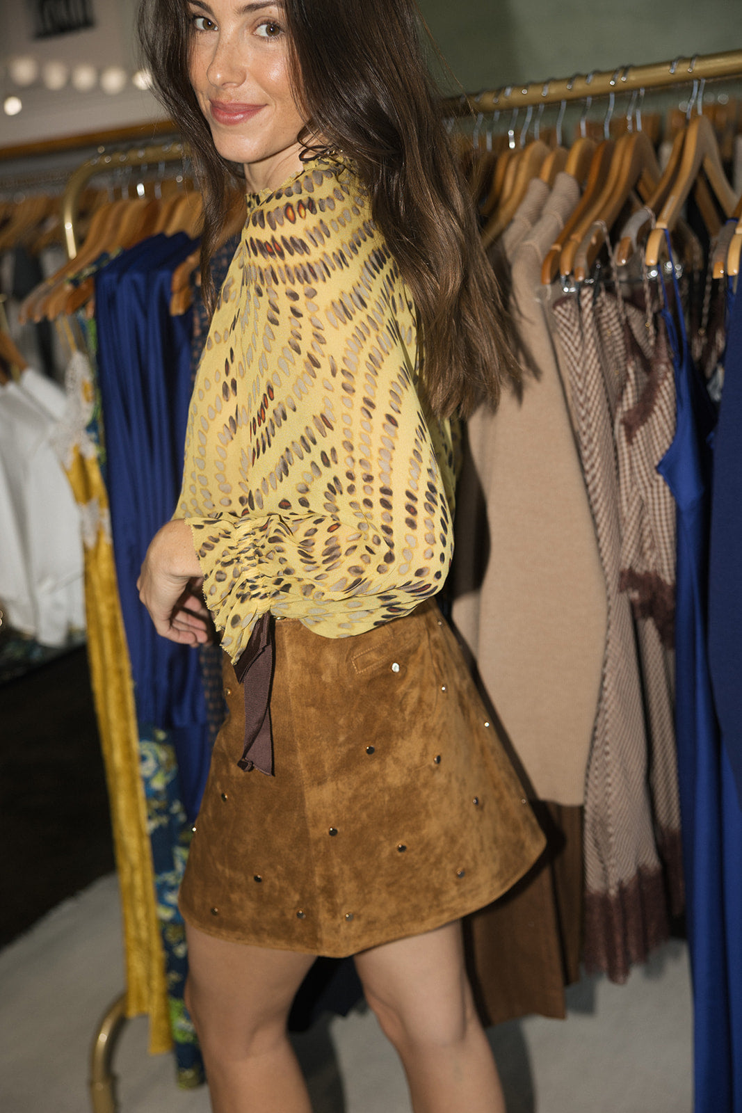 A woman modeling a yellow and brown patterned knit mock-neck top paired with a brown suede mini skirt with rhinestone embellishments, standing in a clothing store with racks of garments in the background.