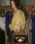 A woman wearing a yellow and brown patterned blouse and brown skirt, carrying a shiny dark brown shoulder bag with a gold-tone crescent moon detail, standing in front of a clothing rack.