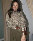 A woman wearing a beige and brown leopard-print long-sleeve blouse with rolled cuffs and an olive green skirt, standing in a fitting room with beige curtains.