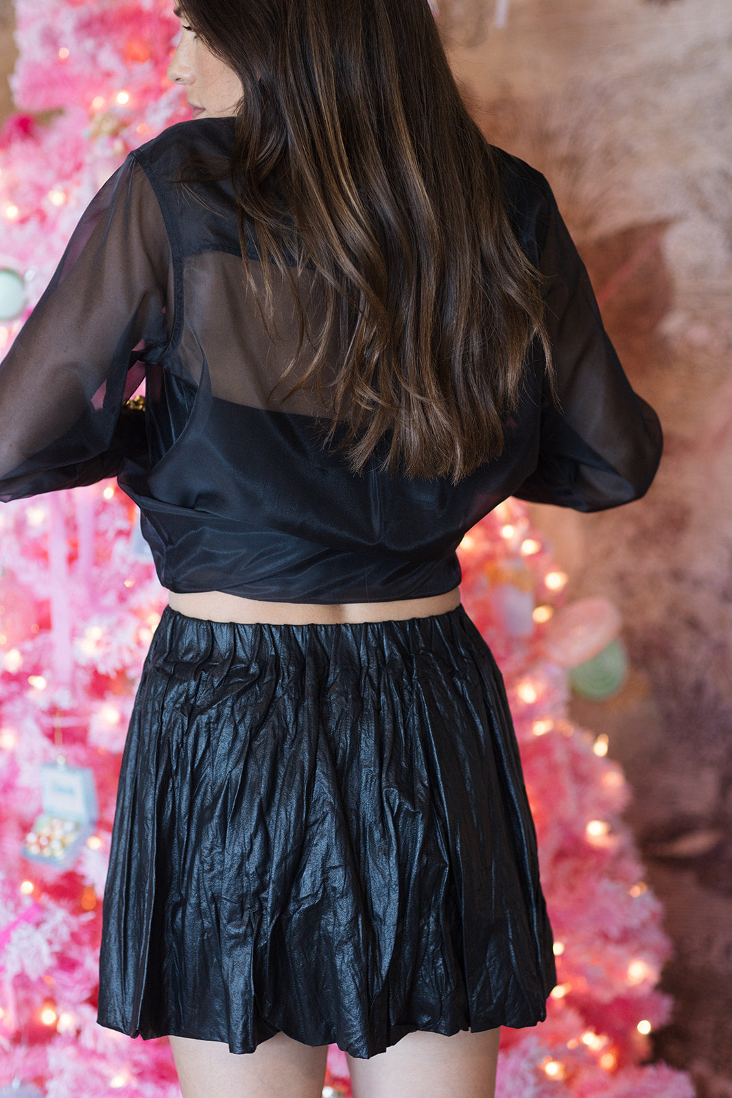 Person wearing a black outfit with sheer sleeves in front of a pink decorative background, back view