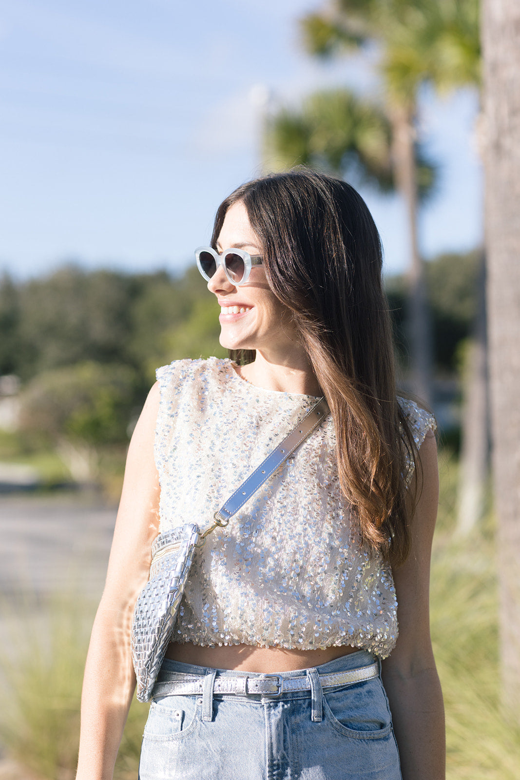 Woman wearing a white sequin top and denim pants outdoors with trees in the background