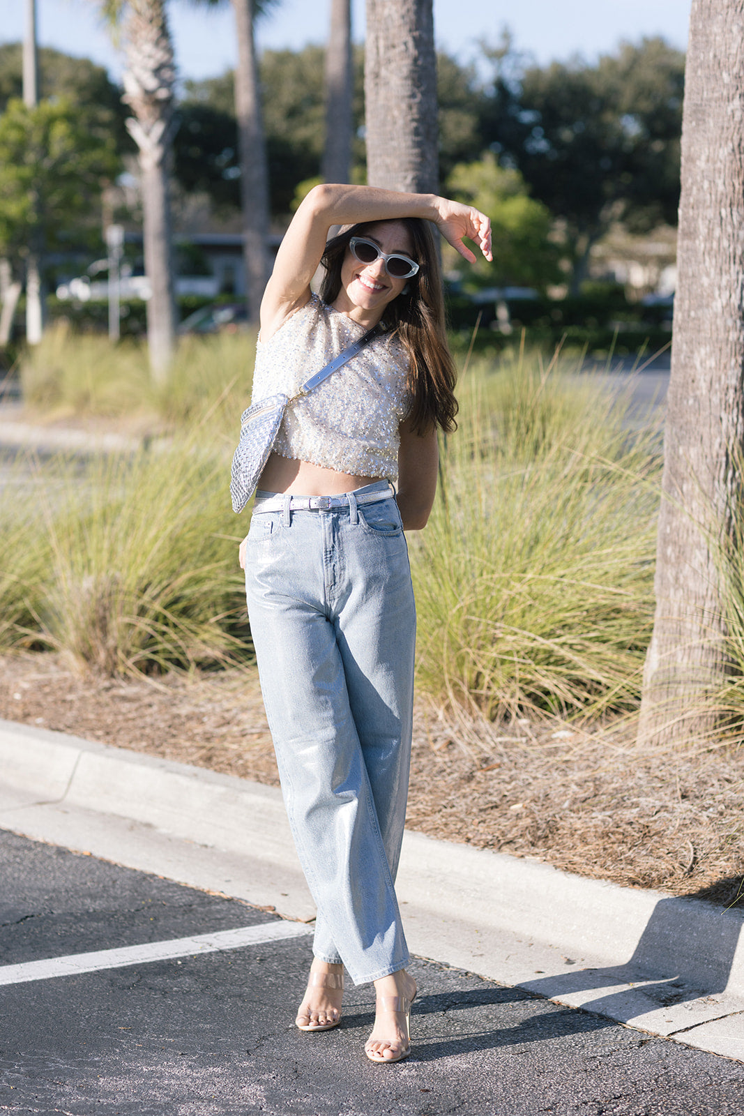 Woman in light blue jeans and a white top standing on a street with palm trees in the background, full body front