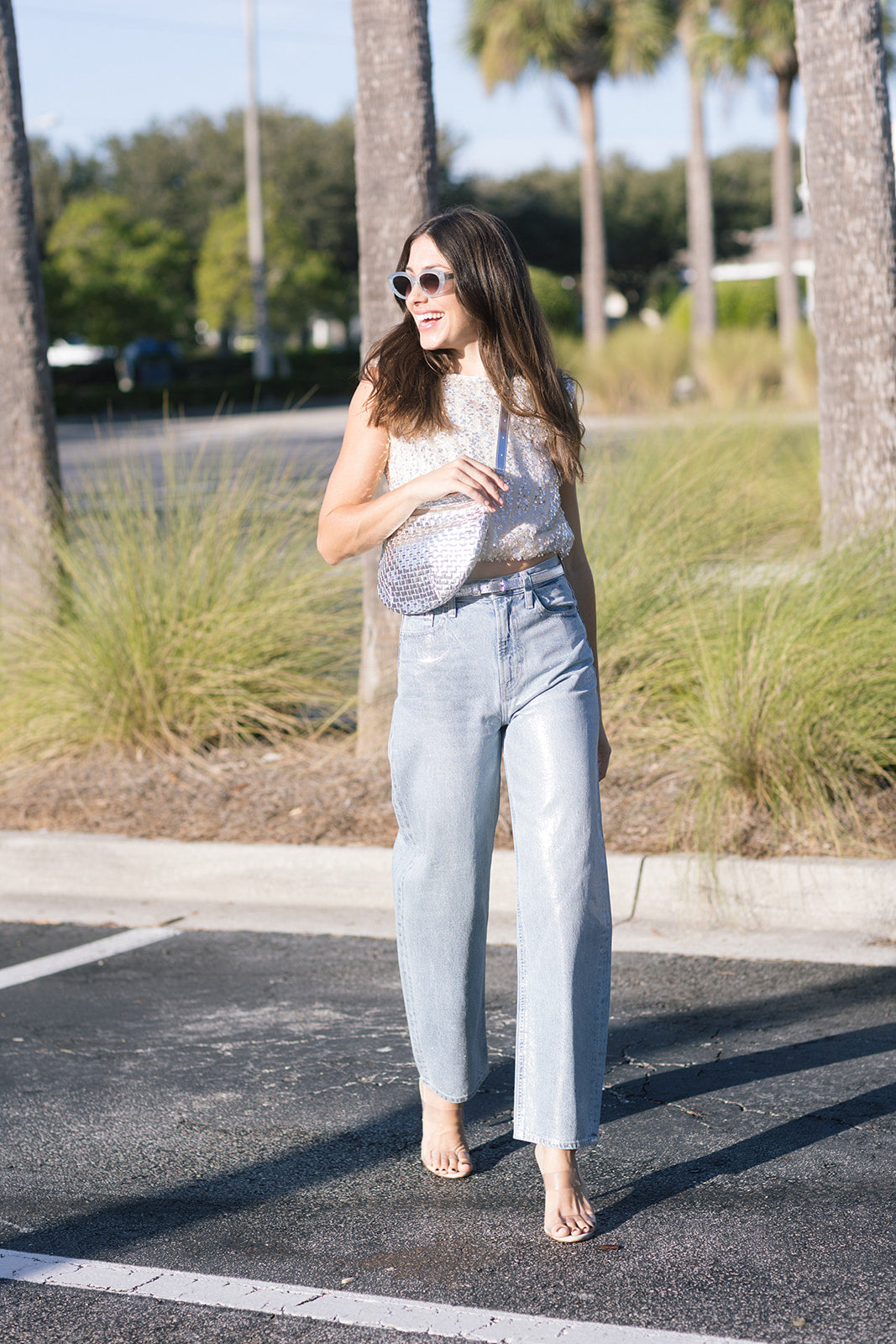 Woman in a sequin top and light blue jeans standing in a parking lot with palm trees in the background.