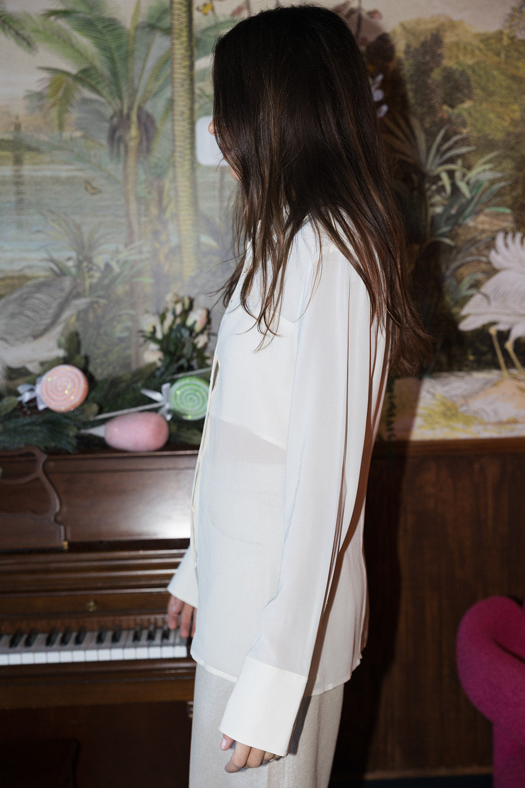Person wearing a white blouse standing in front of a decorative wall with floral patterns. side view
