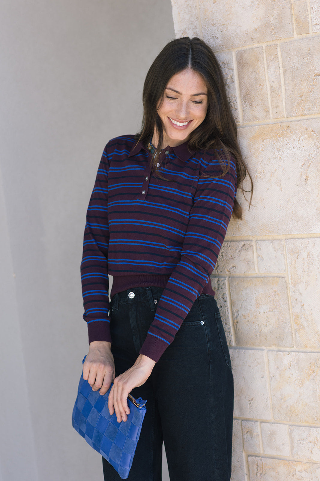 Woman wearing a striped sweater and holding a blue clutch against a stone wall.