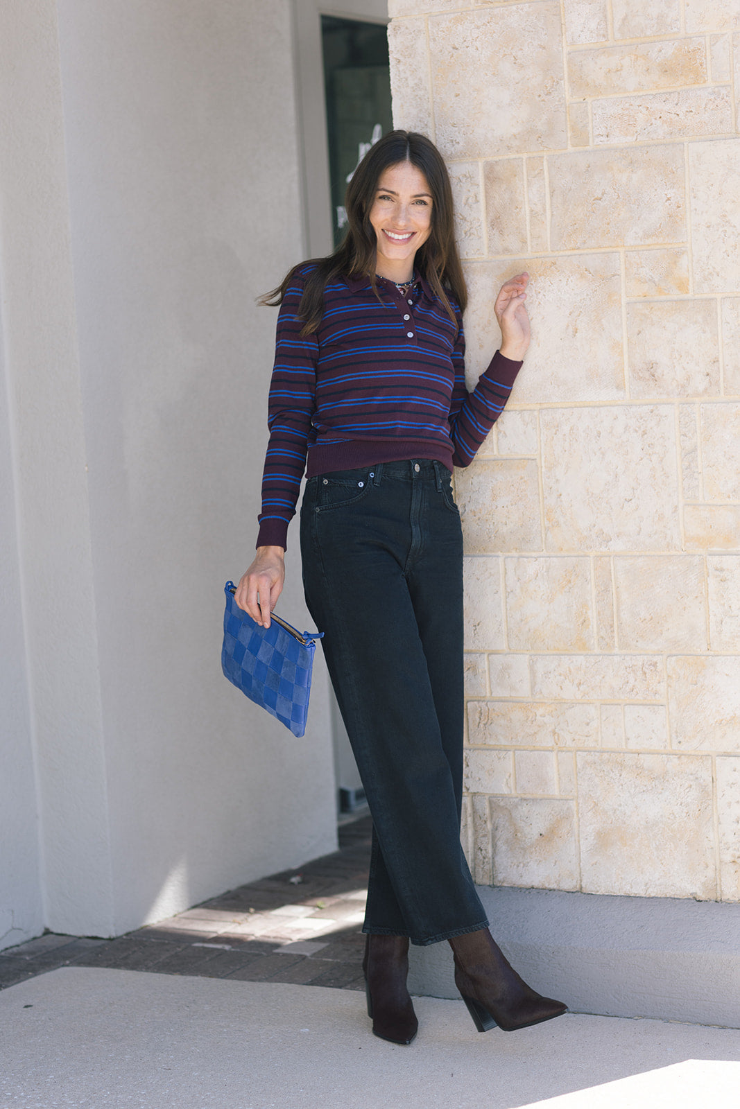 Woman in striped shirt and jeans holding a blue clutch against a stone wall, full body