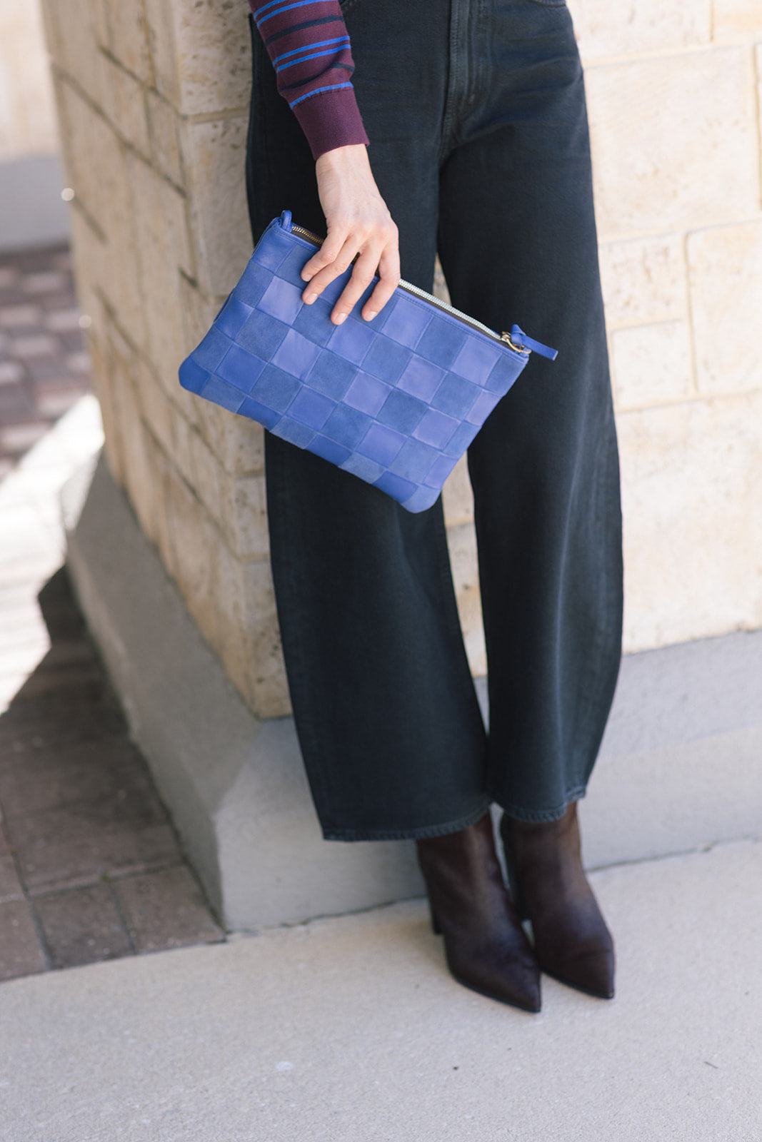 Person wearing black jeans, Mikki Block Heel Boot, holding a blue checkered clutch against a stone wall.