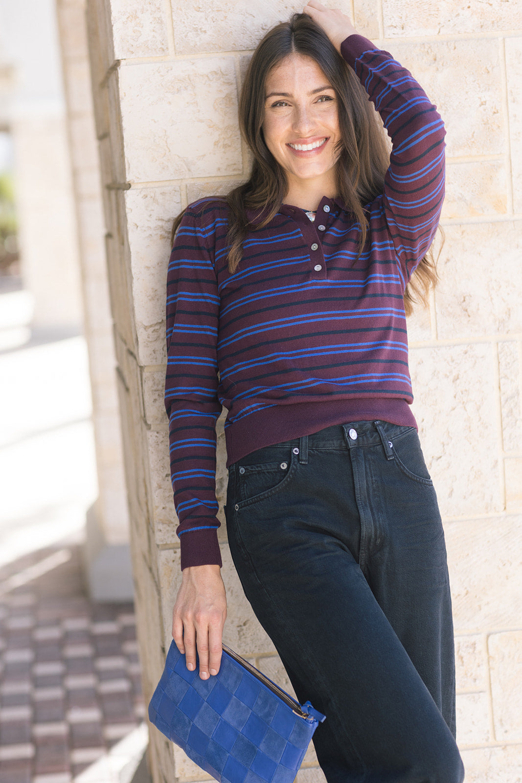 Woman wearing a striped sweater and jeans, holding a blue clutch against a stone wall.