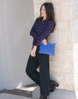 Woman in striped sweater and black pants holding a blue clutch against a stone wall, side view