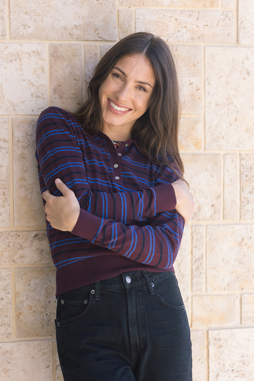 Woman wearing a striped shirt and jeans standing against a stone wall, arms crossed