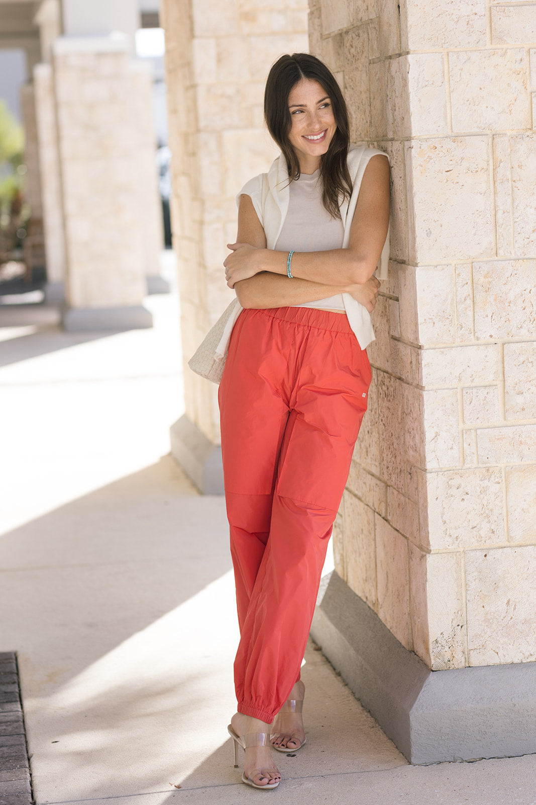 Woman wearing red pants and a beige top standing against a stone wall.
