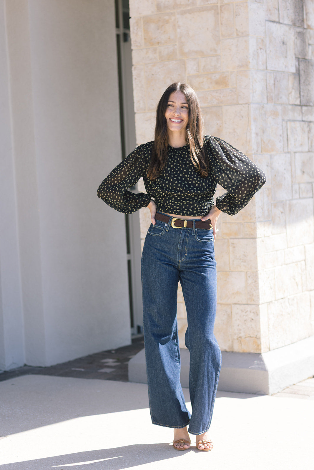 Woman wearing a black polka dot blouse and blue jeans standing outdoors.