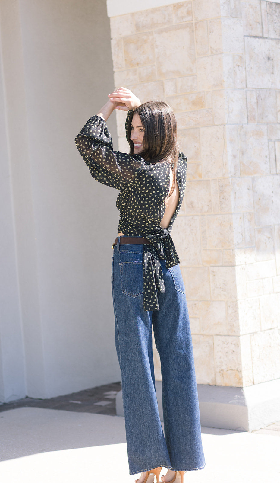 Woman wearing a polka dot blouse and blue jeans against a stone wall, back view
