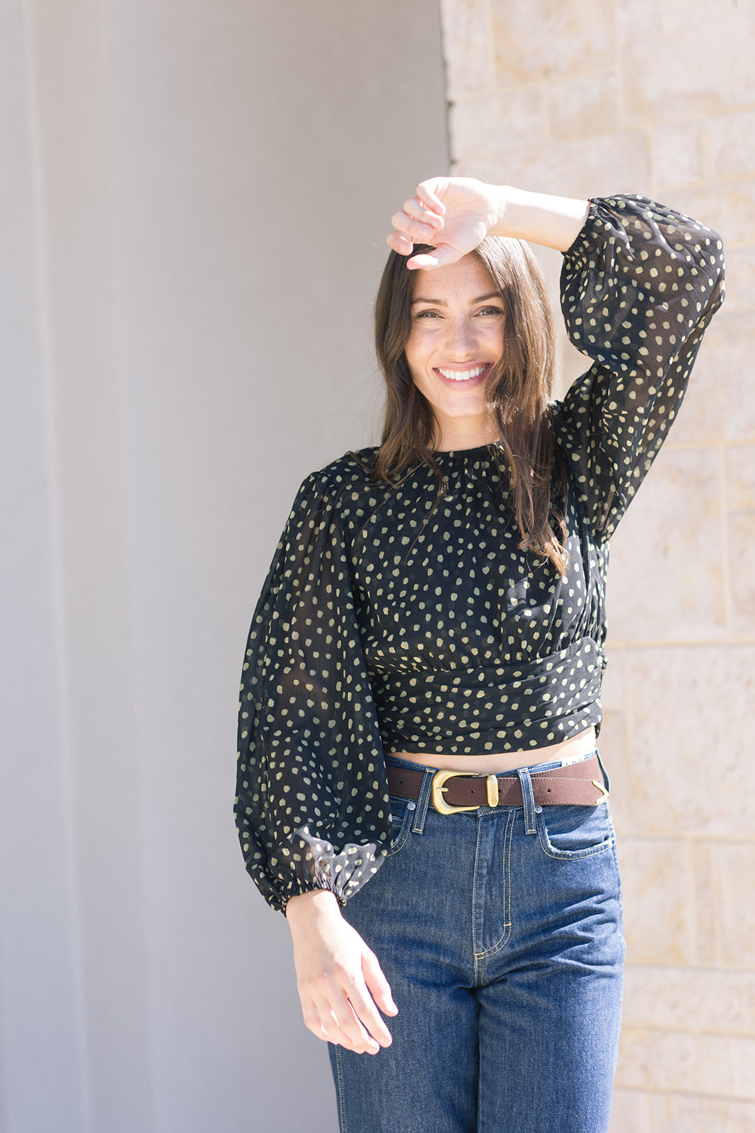 Woman wearing a black polka dot blouse and blue jeans standing against a light-colored wall, top close up