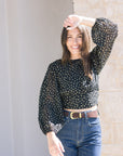 Woman wearing a black polka dot blouse and blue jeans standing against a light-colored wall, top close up
