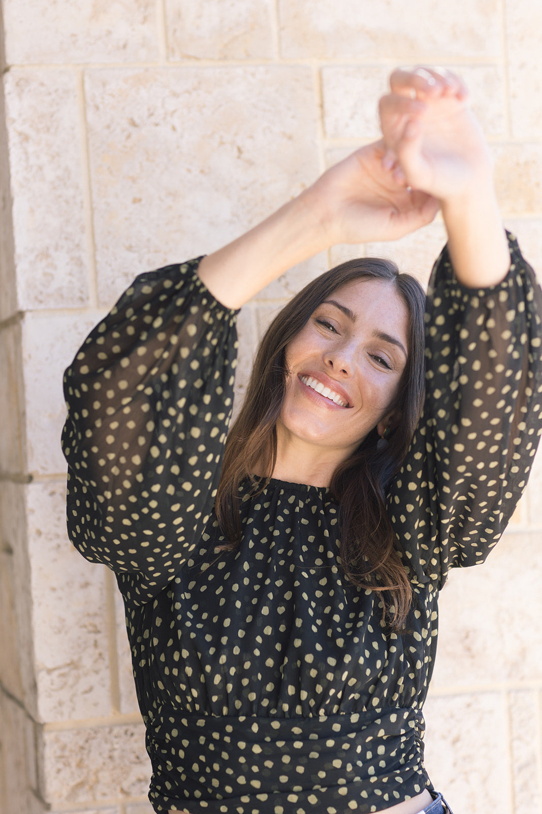 Woman wearing a black polka dot top with arms raised against a stone wall background