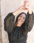 Woman wearing a black polka dot top with arms raised against a stone wall background